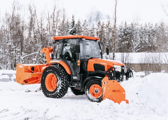 Un tracteur orange équipé d'une fraise à neige déblaie la neige dans une zone enneigée entourée d'arbres et d'un bâtiment. La neige recouvre le sol et les branches, tandis qu'une personne portant un gilet de haute visibilité conduit le tracteur.